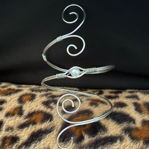 Puede incluir: Un brazalete plateado con un diseño en espiral decorativo. El brazalete presenta un acento de perla central y está hecho de alambre. El diseño incluye espirales decorativas y se muestra sobre un fondo de estampado de leopardo.