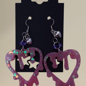 May include: A pair of dangling earrings with a heart shape design. The earrings are a deep pink color with glitter and star accents. The heart shape has a dripping effect. The earrings hang from silver hooks with a small heart charm and purple beads.