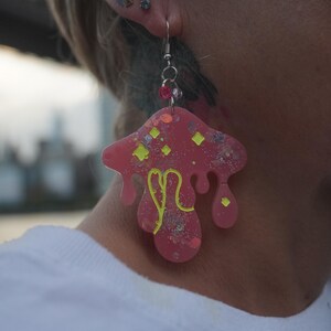 May include: A pink mushroom-shaped earring with yellow and glitter accents. The earring has a silver hook and a small cluster of pink and purple beads. The background is blurred, with a cityscape visible.