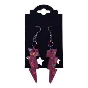 May include: A pair of pink resin lightning bolt earrings with silver-colored hooks. The earrings are filled with glitter and star-shaped confetti. They hang from a black earring display card.