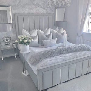 Puede incluir: Un dormitorio lujoso con una cama grande tapizada en tonos grises y blancos. La cama está adornada con numerosas almohadas, una manta suave y una manta blanca texturizada. Una mesita de noche tiene un jarrón con flores blancas. Una lámpara de araña cuelga del techo.