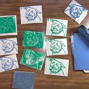 Könnte beinhalten: Eine Sammlung handgefertigter Linolschnittdrucke mit Hundemotiven in verschiedenen Stadien der Fertigstellung. Die Drucke zeigen ein Hundegesicht in grüner und blauer Tinte auf rechteckigem Papier. Ein Farbroller, ein Schneidwerkzeug und ein Farbkissen sind ebenfalls sichtbar.