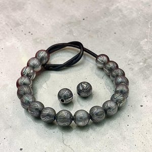May include: A bracelet with gray and white patterned beads, strung on a black cord. Two matching beads are next to the bracelet. The beads have a marbled appearance, creating a unique and stylish accessory.