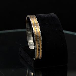 May include: A silver cuff bracelet with two parallel gold-colored stripes. The bracelet is displayed on a black velvet stand against a dark background. The metal has a textured, lined pattern.