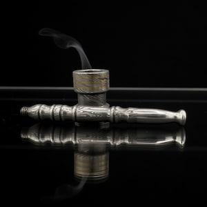 Könnte beinhalten: Eine silberne Rauchpfeife mit gemustertem Design und einem goldenen Band um den Kopf. Rauchschwaden steigen aus dem Kopf vor einem schwarzen Hintergrund auf. Die Pfeife spiegelt sich in der glänzenden Oberfläche darunter.