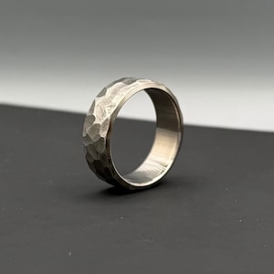 May include: A silver-colored ring with a textured, hammered finish. The ring is round with a smooth interior and a slightly irregular exterior. The ring is set against a dark background, highlighting its metallic sheen.