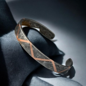 May include: A silver cuff bracelet with a patterned design and copper-colored accents. The bracelet features a geometric pattern and is displayed on a dark blue surface.