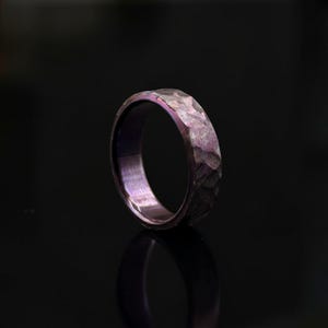 May include: A ring with a dark purple, metallic finish and a hammered, faceted exterior texture. The band features a smooth, polished interior surface that reflects light, creating a deep violet hue across the entire circular metal structure.
