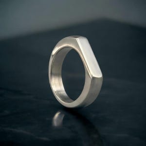 May include: A silver ring with a geometric design. The ring has a flat, rectangular top that tapers to a rounded band. The ring is made of a shiny, reflective metal and is set against a dark background.