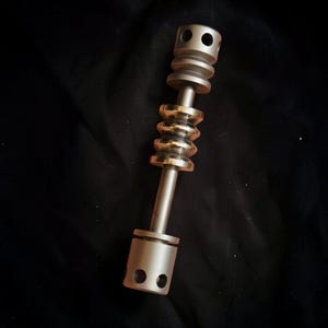 May include: A metallic, cylindrical tool with a series of stacked, ridged sections in gold and silver tones. The tool has a central rod and rounded ends with drilled holes, set against a dark background.