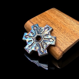 May include: A metallic, snowflake-shaped fidget spinner with an iridescent, heat-anodized finish displaying shades of blue, purple, and gold. The object features a central hexagonal hole and symmetrical, geometric cutouts throughout its six arms.