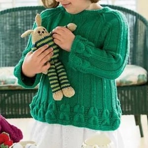 May include: A green knitted sweater with textured details and long sleeves. The sweater is being held by a person. A stuffed bunny with striped accents is held in the person's hands. The sweater has a round neckline and a textured hem.