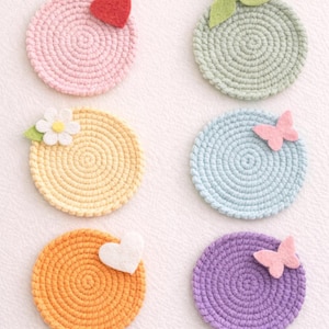 Könnte beinhalten: Sechs farbige gehäkelte Untersetzer in Rosa, Grün, Gelb, Blau, Orange und Lila. Jeder Untersetzer hat ein Spiraldesign und Filzverzierungen wie Herzen, Schmetterlinge und Blumen. Die Untersetzer haben einen Durchmesser von etwa 10 cm.