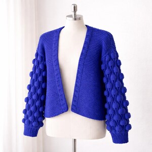 Puede incluir: Un cárdigan azul vibrante con un diseño único. Las mangas presentan un patrón texturizado de punto de burbujas, mientras que el cuerpo tiene un tejido más simple. El suéter abierto se muestra en un maniquí, mostrando su forma y estilo. Una prenda cómoda y elegante.