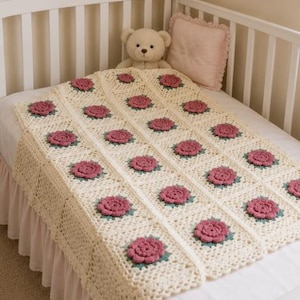 May include: A cream-colored crochet blanket with a floral pattern of pink roses and green leaves. The blanket is draped over a white crib, with a teddy bear and a pink pillow visible in the background. The blanket's edges have a scalloped design.