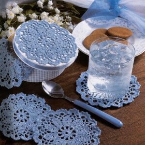 May include: A set of round, light blue crocheted doilies featuring an intricate floral pattern with scalloped edges. These circular textile coasters are displayed alongside a glass of water with ice, a silver spoon, and a plate of cookies.