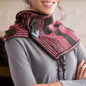 May include: A knitted cowl in pink and gray with large, dark square buttons. The cowl is designed with a textured stitch pattern and is worn around the neck. The color scheme is a mix of pink and gray, creating a visually appealing accessory.