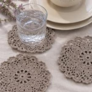 Puede incluir: Tres posavasos redondos de ganchillo en color beige con un intrincado patrón floral calado y bordes festoneados. Un posavasos sostiene un vaso transparente lleno de agua, mientras que los otros dos están colocados por separado.