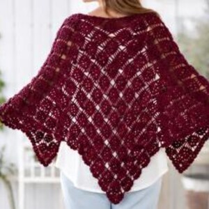 May include: A deep burgundy crochet poncho featuring an intricate diamond-shaped openwork pattern. The garment has a pointed hemline with a scalloped edge, constructed from yarn with a visible texture, draped over a white top and light blue pants.