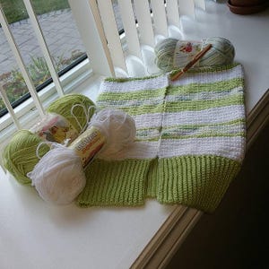 Könnte beinhalten: Ein handgefertigtes Häkelprojekt mit grüner und weißer Wolle. Das Projekt ist ein Kleidungsstück mit grünen Rippenkanten und einem weiß-grünen gestreiften Körper. Mehrere Wollknäuel und eine Häkelnadel sind ebenfalls zu sehen.