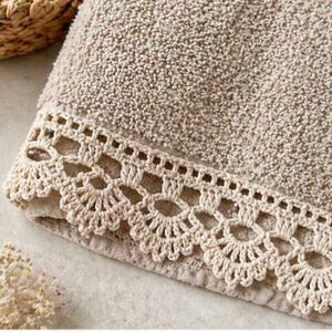 Puede incluir: Toalla de baño beige con un borde decorativo de ganchillo. La toalla tiene una superficie texturizada y un borde doblado. El borde de ganchillo es de un tono beige más claro y presenta un diseño festoneado.