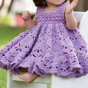 May include: A sleeveless, lavender crochet dress with a square neckline and a full skirt. The dress features an openwork pattern and is designed for a baby or toddler. The child is seated on a white chair, with a blurred green background.