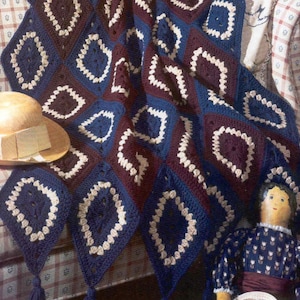 Könnte beinhalten: Handgefertigte Häkeldecke mit geometrischem Rautenmuster in Marineblau, Burgunderrot und Creme. Die Decke ist über eine Oberfläche drapiert, mit einem Strohhut und einer Puppe. Die Decke hat Quasten.