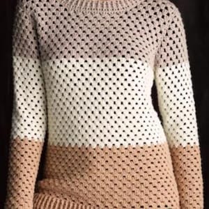 May include: A crochet sweater in a color block design. The top is a light brown, transitioning to white in the middle, and a darker tan at the bottom. The sweater has long sleeves and a crew neck.