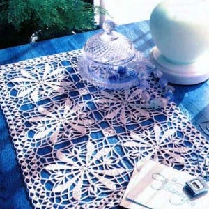 May include: A white, open-weave crochet table runner with a floral design, resting on a blue surface. A clear glass dish with a lid and a white teacup are also visible. A small card with the word "Thank You" is on the right.