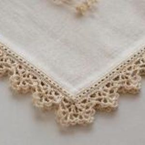 May include: A close-up shot of a white linen handkerchief featuring a cream-coloured, crocheted lace border. The lace trim provides a vintage, handcrafted appearance to the square cloth.