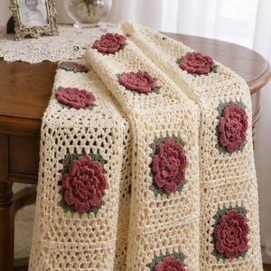 May include: Cream-colored crocheted blanket with square floral patterns. Each square features a burgundy flower with green leaves. The blanket is draped over a wooden table, showcasing its handmade texture and design.