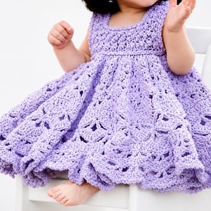 Puede incluir: Un vestido de ganchillo lila con cuello cuadrado y falda acampanada. El vestido presenta un patrón calado detallado y está diseñado para una niña. El vestido está hecho a mano y parece suave y cómodo.
