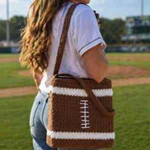 May include: A brown and white crochet crossbody bag designed to resemble a football. The bag has a rectangular shape with white stripes and a white stitched design. The strap is brown and appears to be crocheted as well.