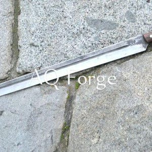 Peut inclure: Une longue épée argentée avec un manche en bois et une extrémité incurvée. La lame est large et plate, avec un bord tranchant. Le texte "AQ Forge" est visible sur la lame.