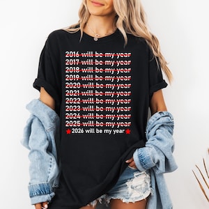 May include: Black t-shirt with the text "2016 will be my year" through "2026 will be my year" in white font, with red lines through the years except for 2026. A red star is next to the year 2026.