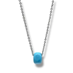 Può includere: Collana con catena d'argento e pendente azzurro a forma di barilotto. Il pendente è di colore pieno con finitura liscia e lucida. La catena è delicata e pende dal pendente.