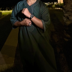 May include: A dark teal tunic with embroidered detailing around the collar and sleeves. The tunic is worn with a watch with a black face and a gold band. The person is also wearing a ring. The background is dark with a path and trees.
