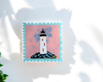 Może przedstawiać: Kwadratowa naklejka z ilustracją latarni morskiej. Biała latarnia morska z czarnym dachem i czerwonymi akcentami na tle różowo-niebieskiego nieba. Naklejka ma jasnoniebieską obwódkę z motywem znaczka pocztowego.