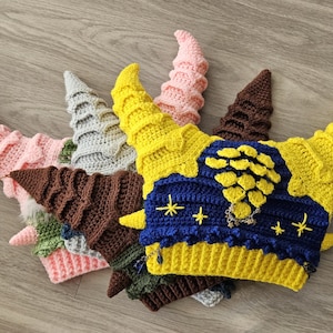 Può includere: Una collezione di cappelli all'uncinetto fatti a mano in vari colori, tra cui rosa, grigio, marrone, giallo e blu. I cappelli presentano un design a forma di corno e sono disposti su una superficie di legno. Alcuni cappelli hanno dettagli di stelle e catene.