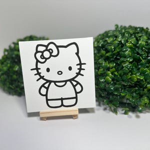 Puede incluir: Un lienzo cuadrado blanco con un contorno negro de Hello Kitty, presentado sobre un pequeño caballete de madera. El lienzo está centrado, con dos plantas decorativas verdes y esféricas en el fondo.
