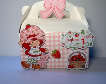 May include: A white gift box with a pink bow, featuring a Strawberry Shortcake character and a pink and white checkered pattern. The box is decorated with strawberry and basket graphics, and the text "Jaylenis 1 Cumple".