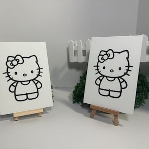 Puede incluir: Dos lienzos blancos con un contorno negro de Hello Kitty. Cada lienzo está apoyado en un pequeño caballete de madera. La obra de arte está sobre un fondo blanco con arbustos verdes y una valla blanca.