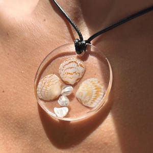 May include: A clear, circular pendant necklace featuring three seashells with orange and white patterns, and small, clear stones. The pendant is suspended from a black cord.