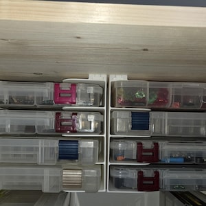May include: Clear plastic storage containers with colored latches, arranged in a cabinet. The latches are various colors, including pink, burgundy, and blue. The containers are filled with small items, likely craft supplies or hardware.