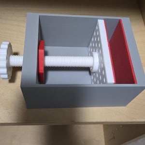May include: A gray rectangular clamp with a white threaded rod and a white knob. The clamp has a red disc and a perforated white panel with a red edge. The clamp is on a wooden surface.