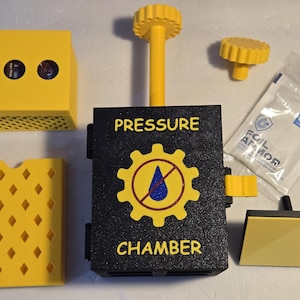 May include: A disassembled pressure chamber kit with yellow and black components. The main black chamber has the words "PRESSURE CHAMBER" and a gear symbol. Additional parts include a yellow cube, a perforated yellow panel, and a small packet.