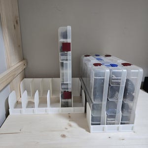 May include: Clear plastic storage containers with red and blue accents, filled with various items. A white organizer with dividers sits to the left. The containers are on a light wood surface.