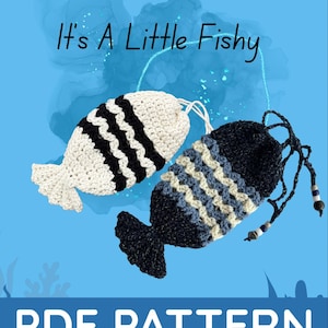 May include: Two crocheted fish-shaped pouches, one white with black stripes, the other navy blue with blue and cream stripes. The text "It's A Little Fishy" and "PDF PATTERN" are displayed on a blue background. A logo with a cat is in the upper right corner.