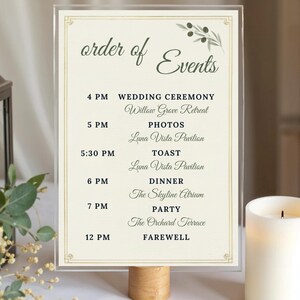 May include: A clear acrylic wedding event schedule with the words "order of Events" in elegant script. The schedule lists times and locations for the ceremony, photos, toast, dinner, party, and farewell. The design includes olive branch illustrations and a wooden base.