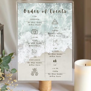 May include: A clear acrylic wedding sign featuring a beach-themed design. The sign details the "Order of Events" with times and locations for the ceremony, photos, toast, first dance, dinner, and party. It is supported by a wooden base.